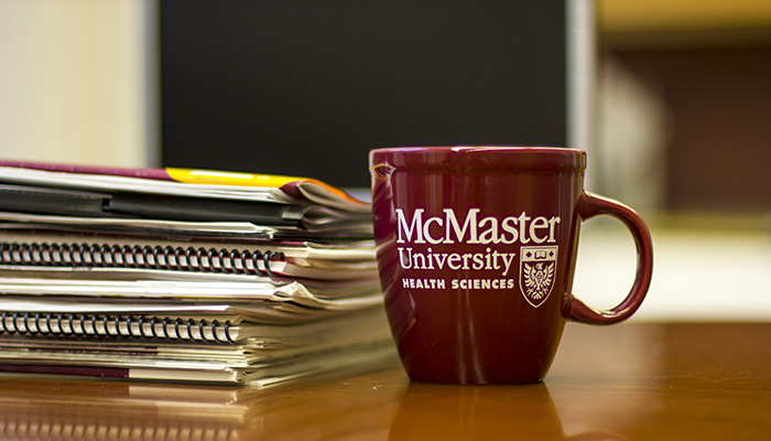 McMaster Health science brand coffee cup on a desk with a stack of papers and books